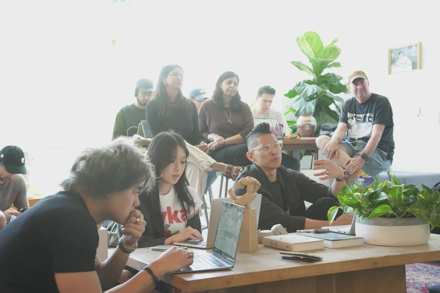A long table of people working on laptops, with more people gathered behind them.